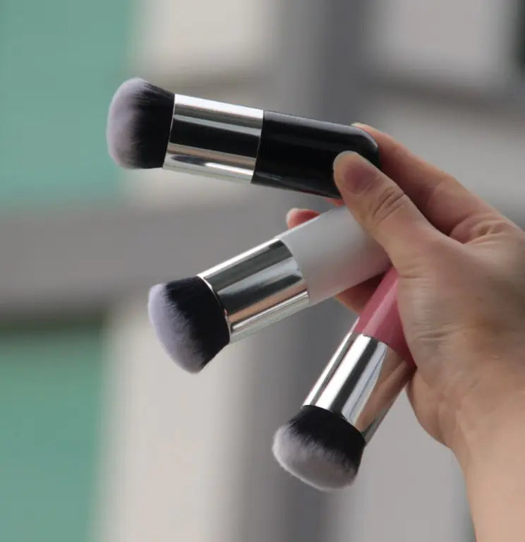 Hand holding three Flawless Finish Foundation Brushes with elegant metal handles in different colors.